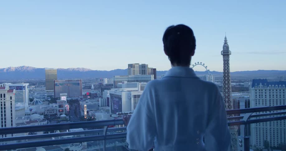 Woman in a white bathrobe leans against the balcony rail as she looks at the city below