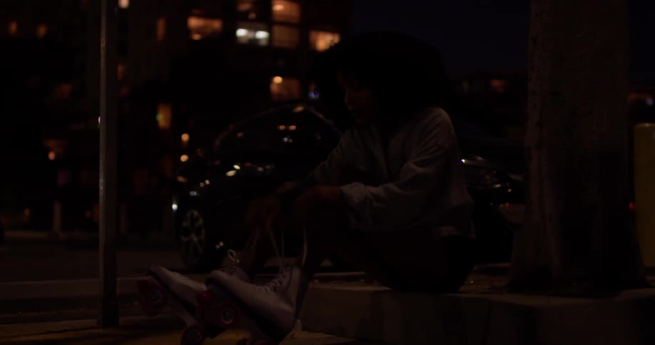 Woman sits outside in the city at night tying her roller skates