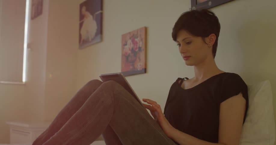 Woman sits on a bed looking at a tablet