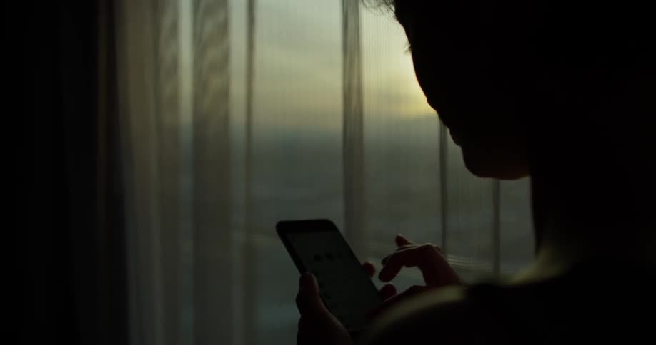 A woman uses her phone next to a window at sunset