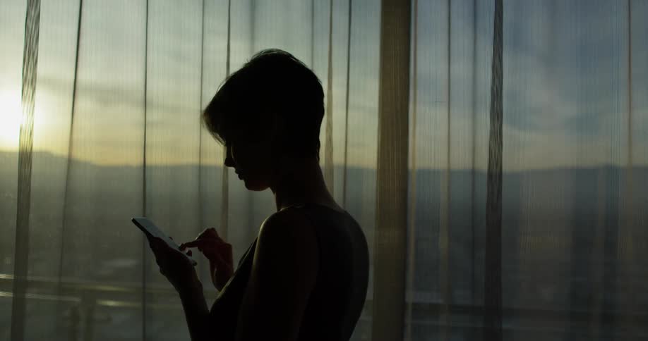 Woman taps her cell phone screen as she stands near a window