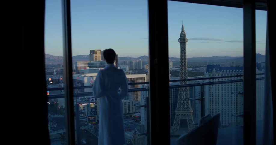 Woman in a white bathrobe stands on a balcony and looks out at the city