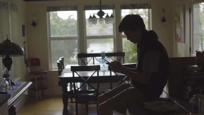 A teenage boy uses a phone while sitting on a kitchen counter