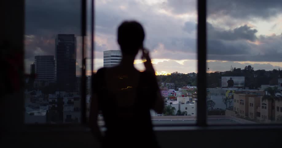 A woman talks on a phone next to a window at sunset