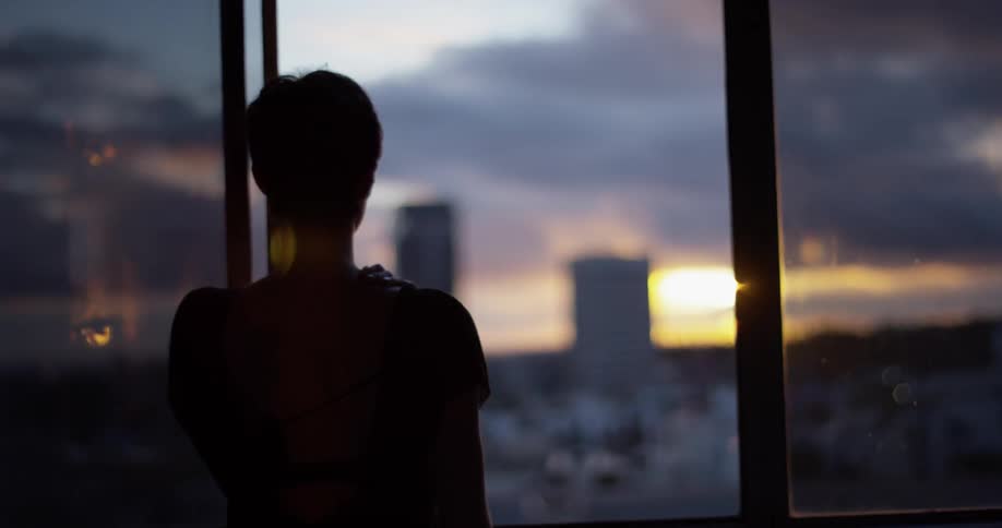 A woman looks out a window at sunset