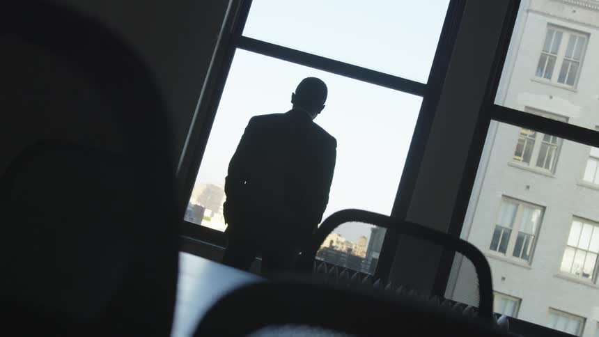 A man in a suit stands in a conference room and looks out the window