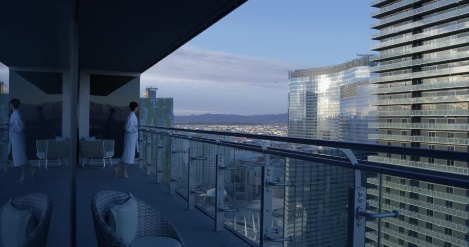 Woman in a white bathrobe stands on a balcony and looks out at the city