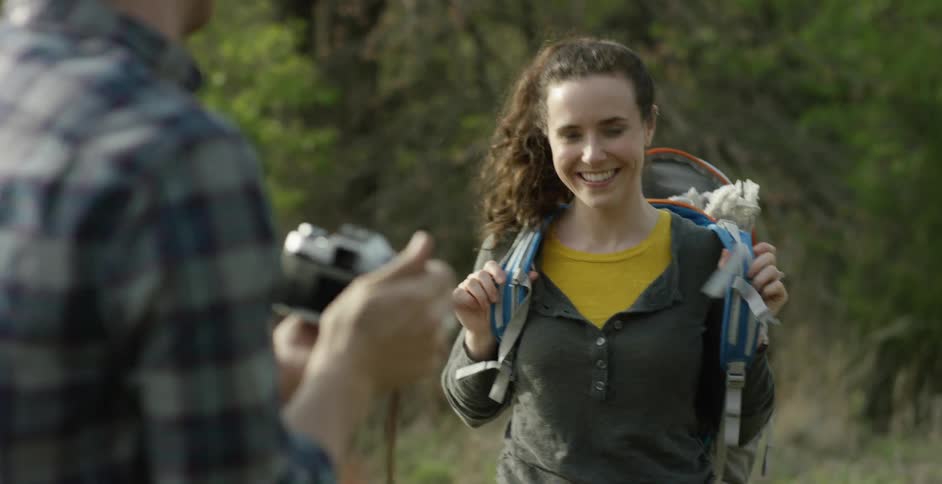 A brunette woman putting on a backpack and smiling at a brunette man who is photographing her
