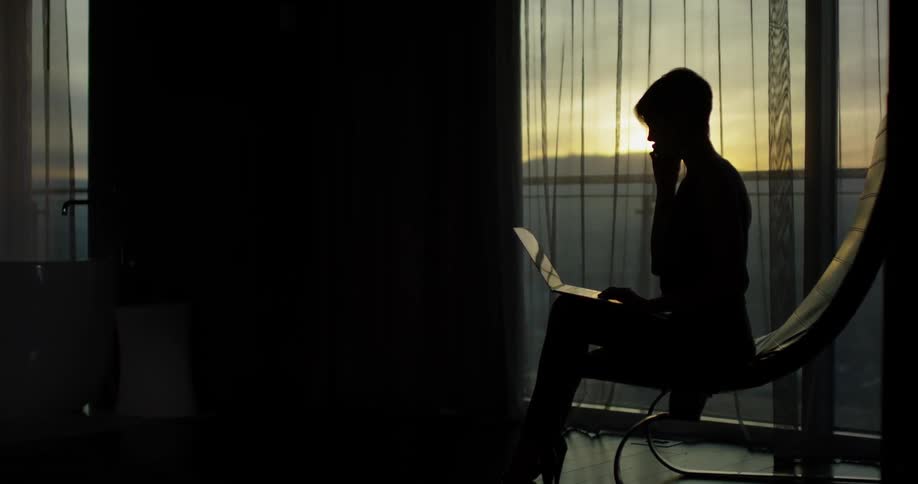 A woman is sitting with her laptop and phone in the dark room