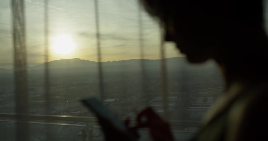 A woman uses her phone next to a window at sunset