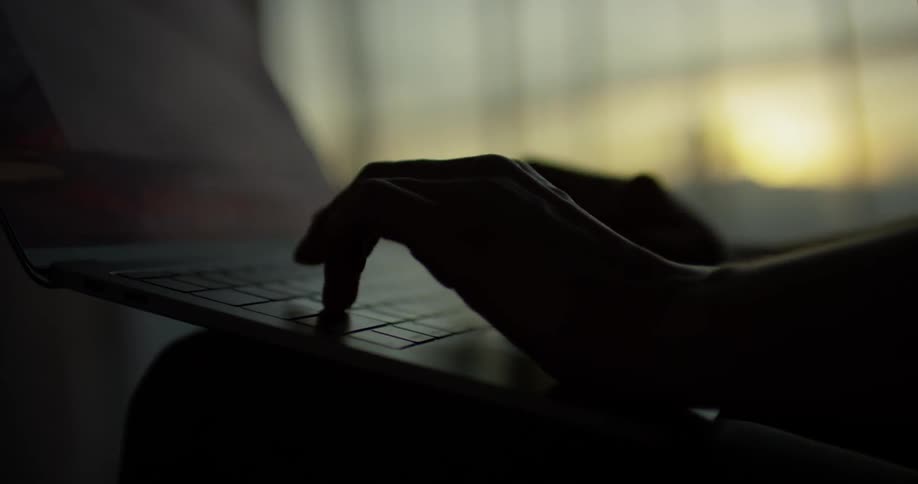 Person's hands type on a laptop next to a window at sunset