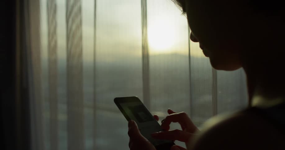 A woman uses her phone next to a window at sunset