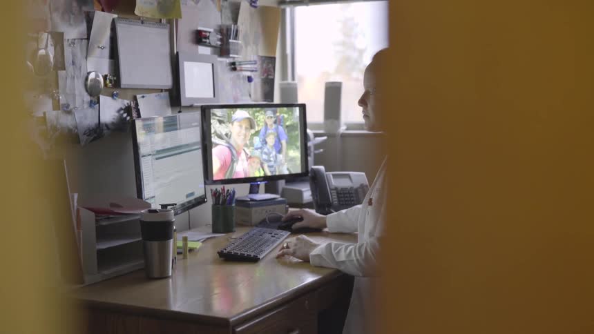 A woman in a white lab coat sits at a desk in an office working on a computer