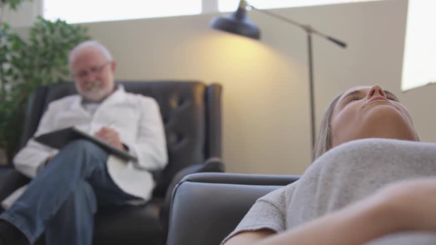 A woman is lying on a therapist's couch as he listens to her