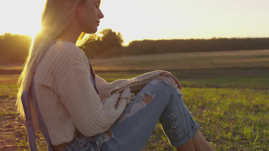 Woman is sitting in grass field at sunset