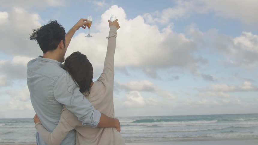 Man and woman stand on the beach raising glasses in the air