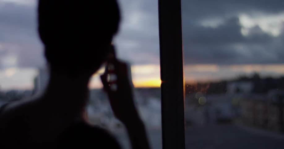 A woman talks on a phone next to a window at sunset