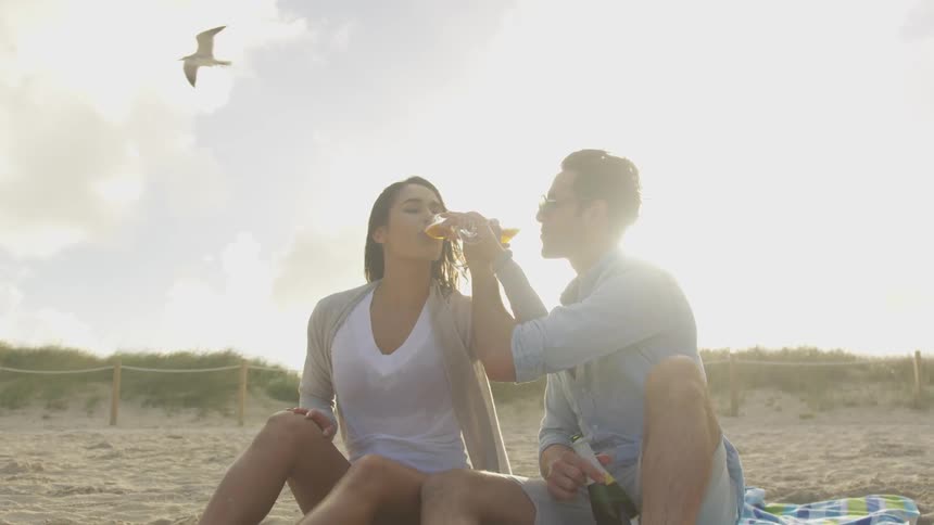 A couple drinks from glasses with entwined arms at the beach