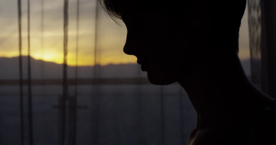 Woman sits next to a large curtained window at sunset