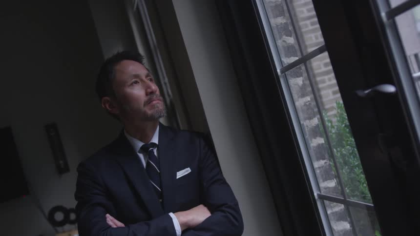 Man wearing a suit stands in a dim room looking out the window