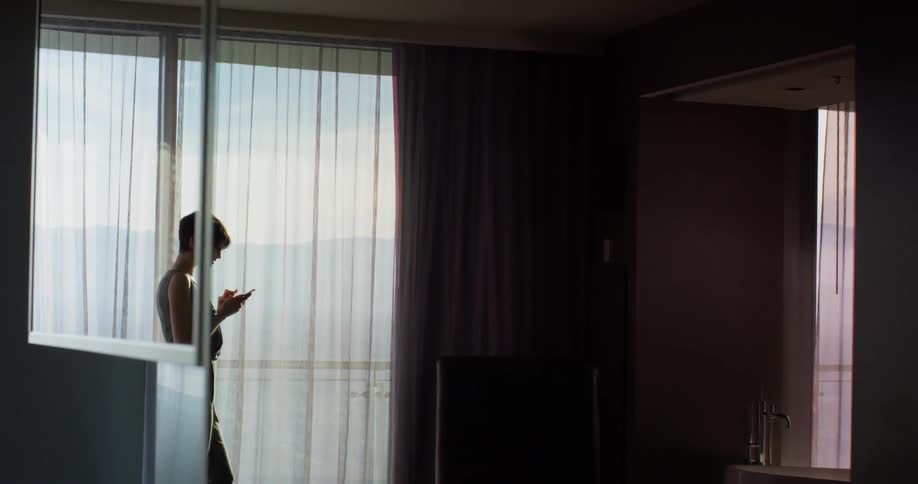 Woman stands near a curtained window as she looks at her cellphone