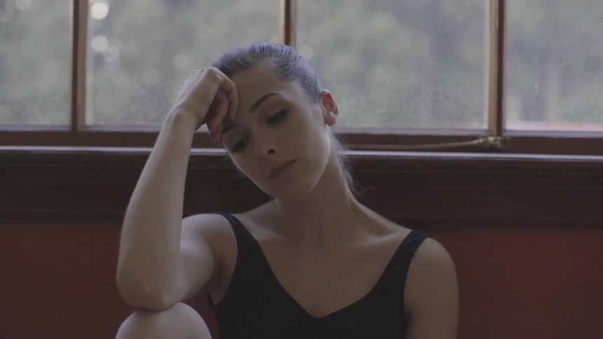 A woman sits in front of a window resting her head on her hand and closing her eyes