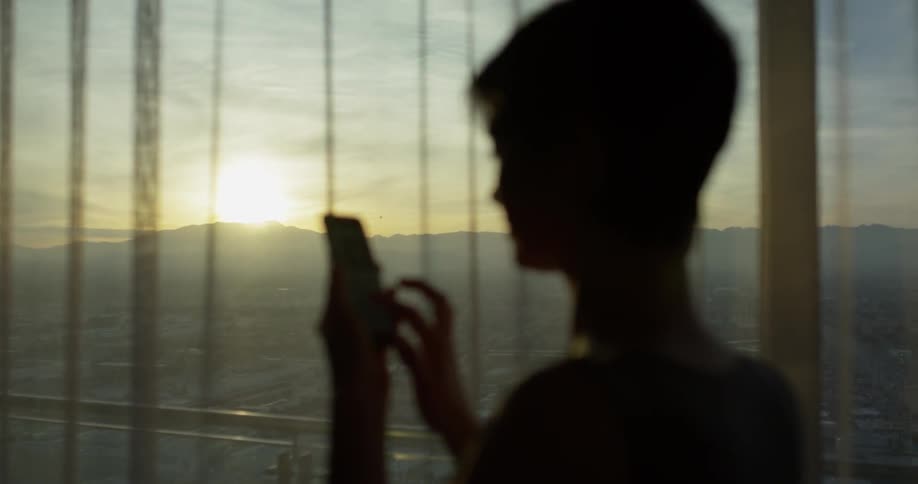 Woman types on her cell phone as she stands near the curtained window