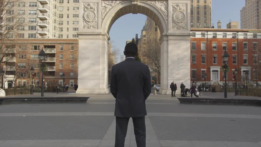 A man stands in a city plaza looking at an arch