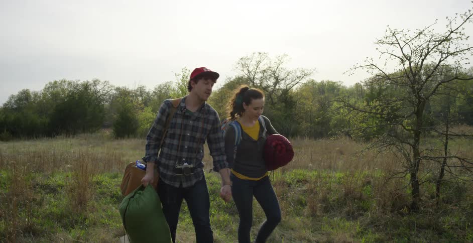 A man and woman carrying camping equipment walk through an open field