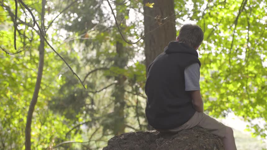 A teenage boy sits on a rock in a forest