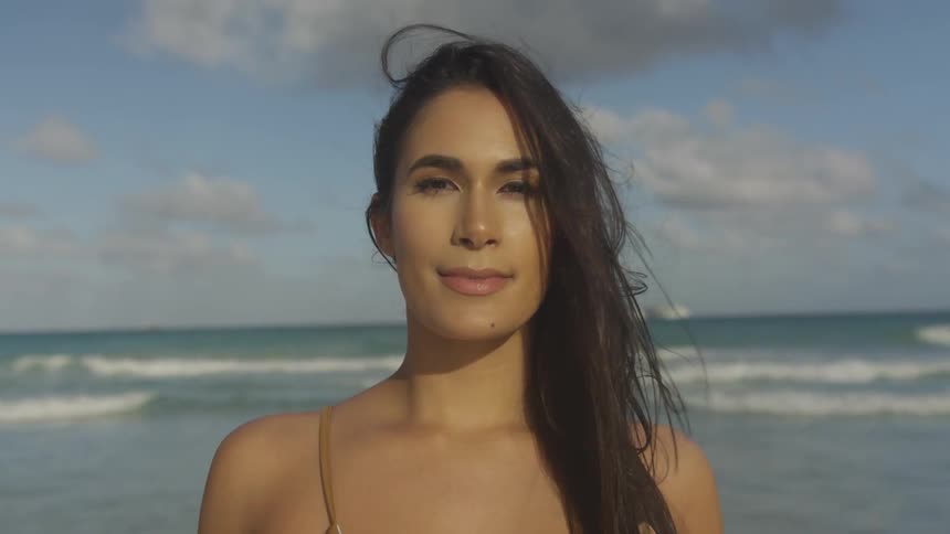 A woman standing on a beach in a bathing suit looks up and smiles
