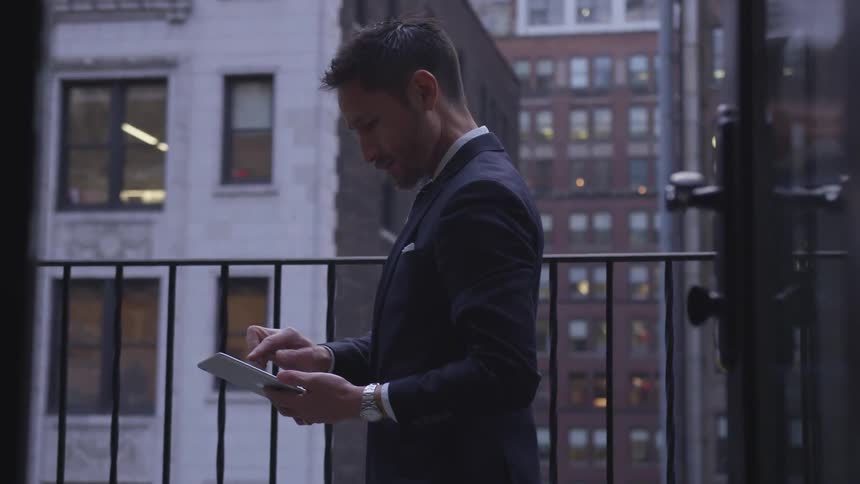 Man types on a tablet while standing on a balcony in the city