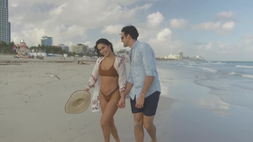 A man bends in to kiss a woman as they walk on a beach in daylight
