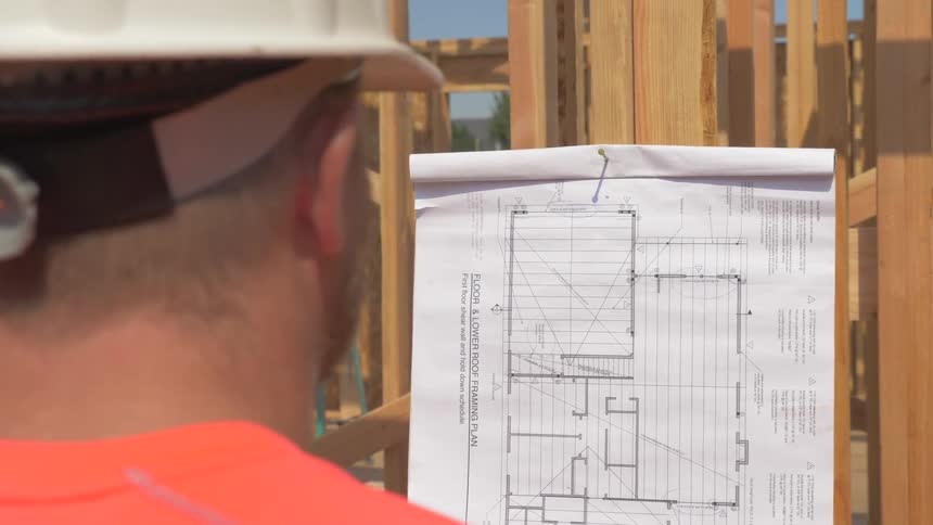 A construction worker is reading blueprints posted on a wall outside in daylight