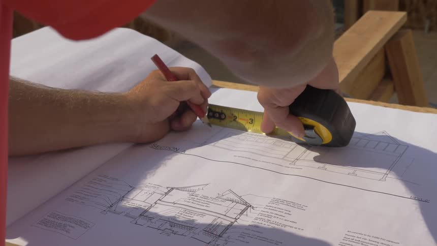 A man is using a measuring tape as he writes on blueprints in daylight