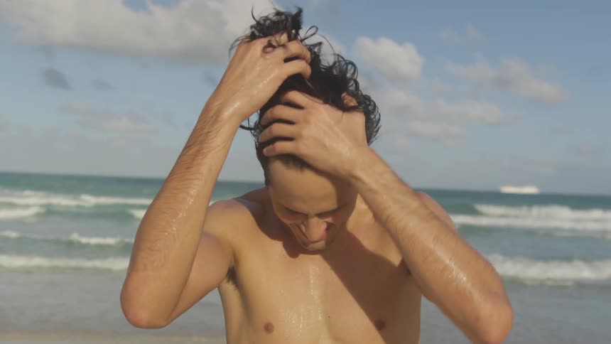 A man stands on the beach on a partly cloudy day running his fingers through his wet hair