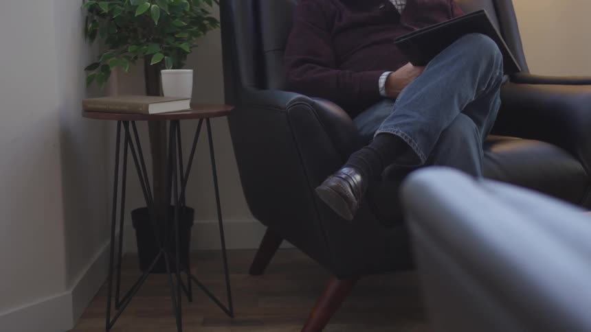 A man sits cross-legged holding a book next to a table with a mug, book and plant on it