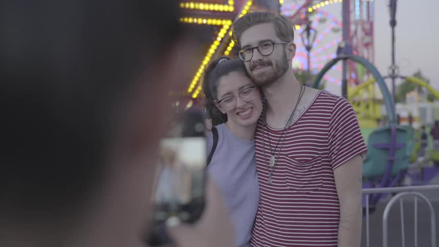 A woman is taking a photo of a couple at a fair at dusk