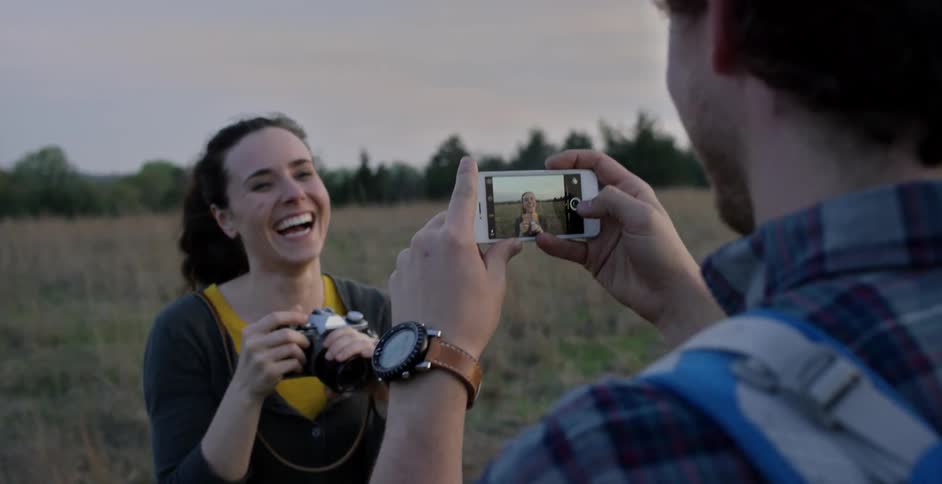 A man and woman stand in an open field laughing and taking pictures of each other