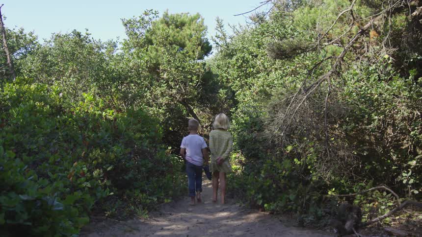 A boy and girl walk on a dirt path through shrubs and trees