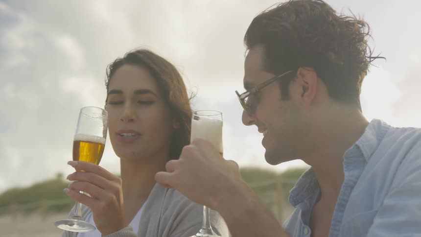 A man and woman drink beer from champagne flutes on the beach