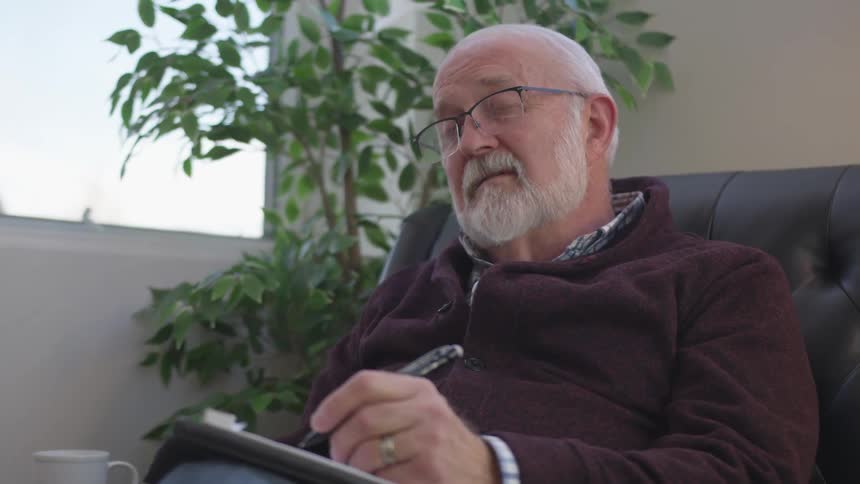 Older man is sitting on chair and writing on notepad