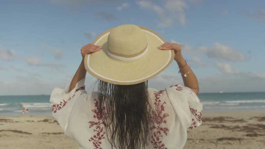 A woman puts on her hat on the windy beach