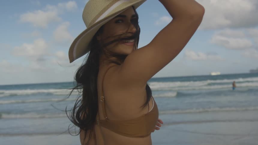 A woman stands on the beach holding her hat as she spins around and smiles