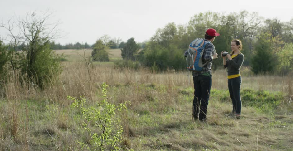 A man and woman walking on grass, the man takes the backpack from the woman