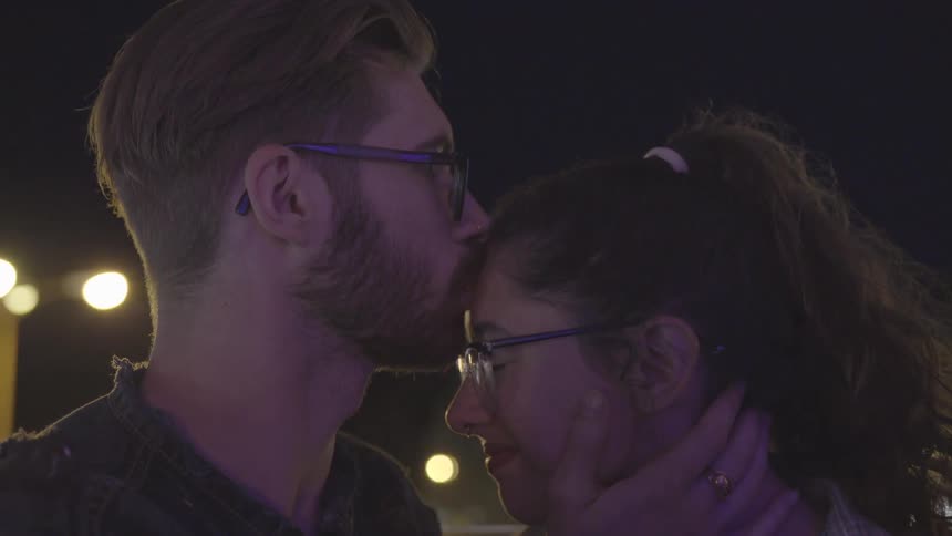 A man holds a woman's face and kisses her forehead as they sit on a ride at night