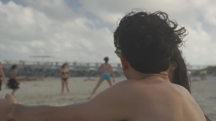 A man and a woman sit on the beach watching a group of people playing volleyball as the man pretends to hit a ball