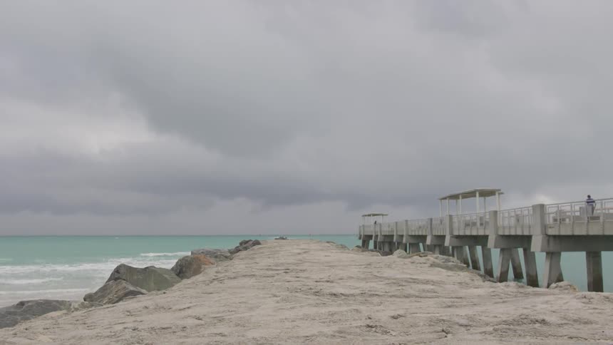 A man walks down a dock by the ocean on a very cloudy day