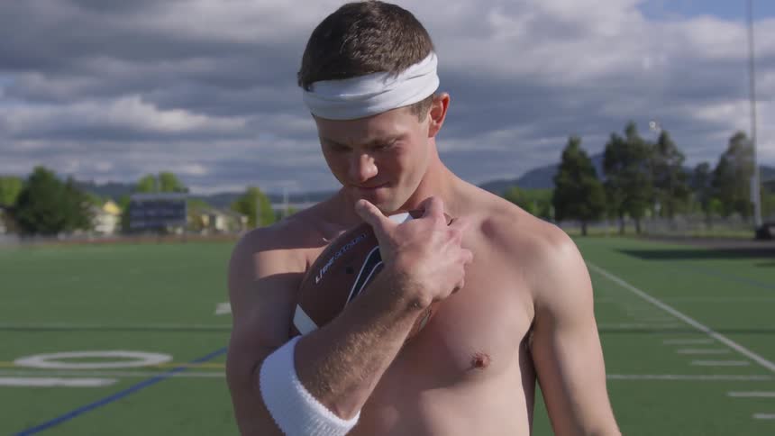 Shirtless man stands in a playing field holding a football