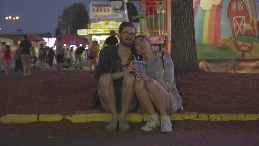 A man and woman sit on the curb by the carnival hugging